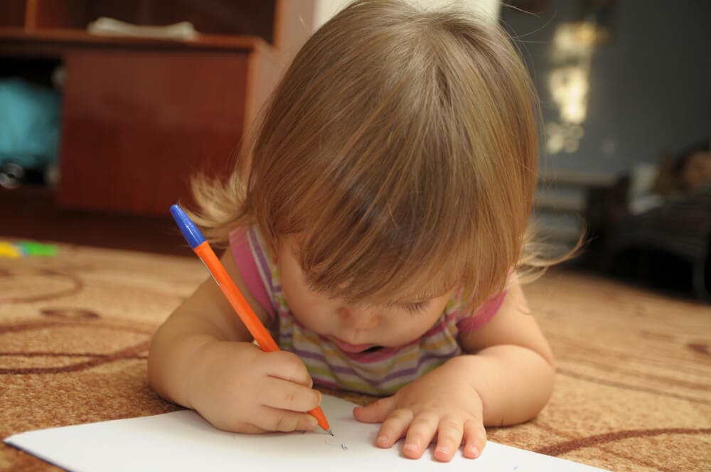 Терпение для ребенка. Терпение для детей учиться писать. Малыш учится писать каракули. Окна развития ребенка. Девочка с лежит на ковре и пишет.